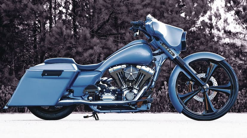Blue custom bagger motorcycle with large black spoked wheels parked on a pavement before dark pine trees.