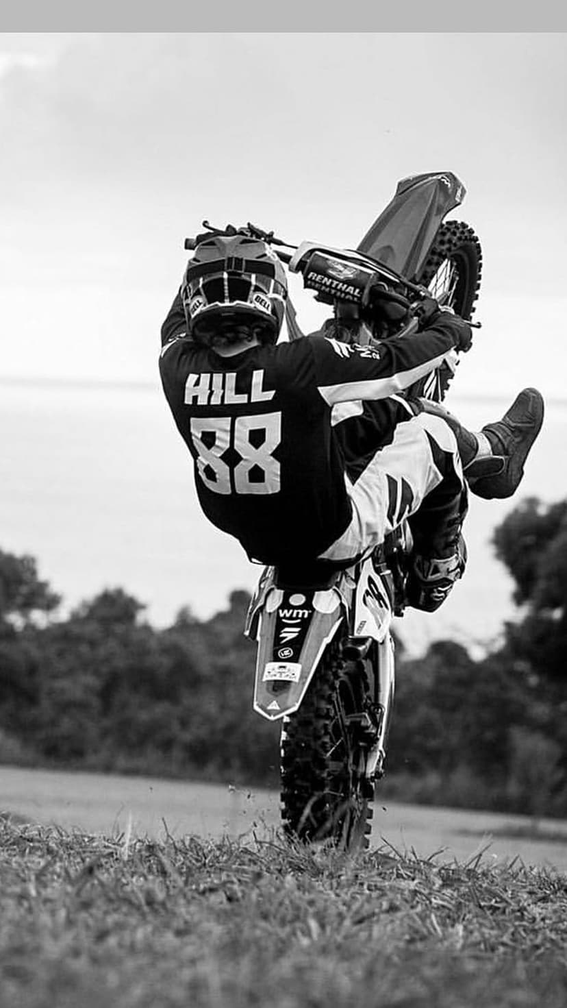 Motocross rider number 88 performs a wheelie on a dirt bike in a grassy field with a misty forest background.