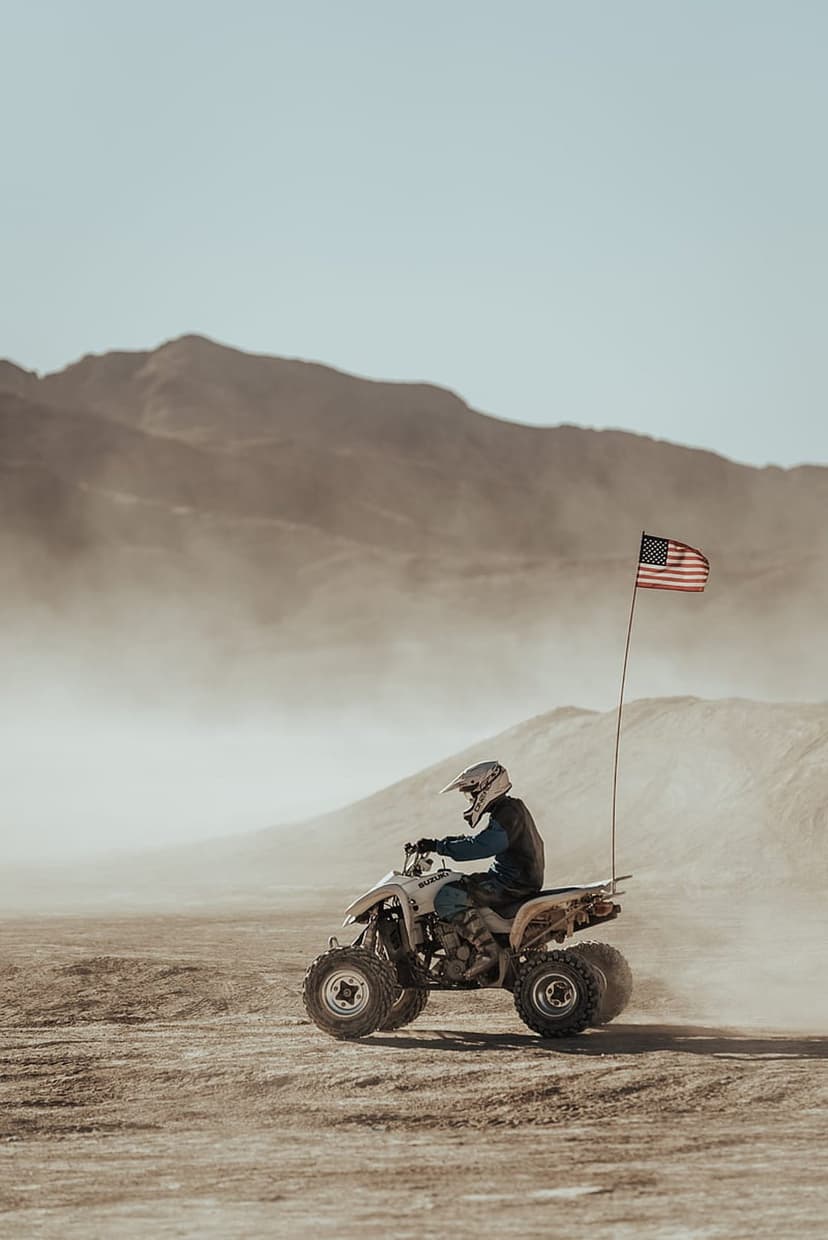 Quad bike rider with a trailing American flag kicking up dust across sunlit, arid desert terrain.
