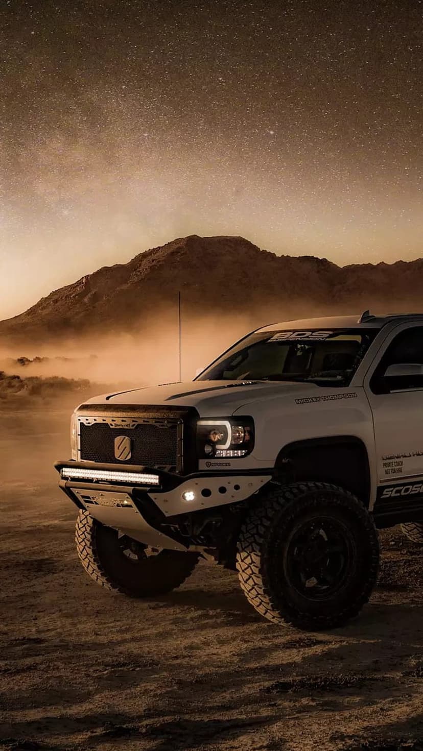 White off-road truck with LED lights parked in a sandy desert under a vibrant, star-filled Milky Way night sky.