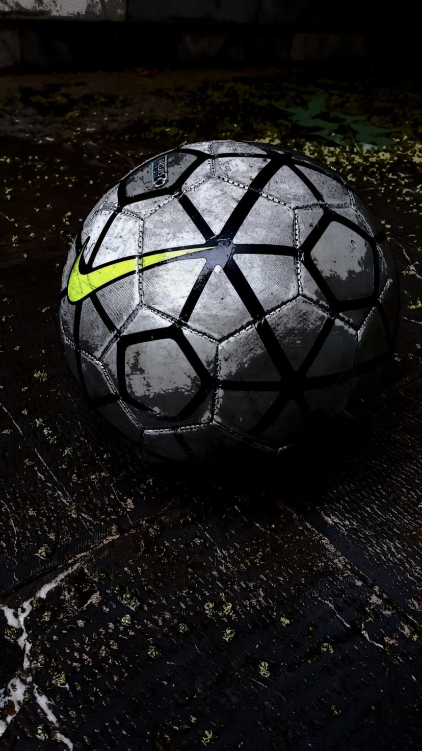 Silver soccer ball with a neon yellow Nike swoosh rests on a dark wet surface with dramatic shadows and debris.