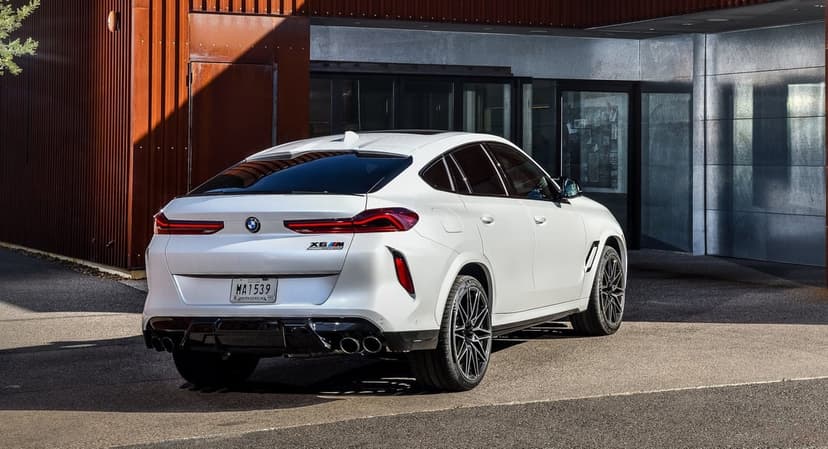 White BMW X6 M Competition rear view with quad exhausts parked in front of modern metallic and rusted buildings.
