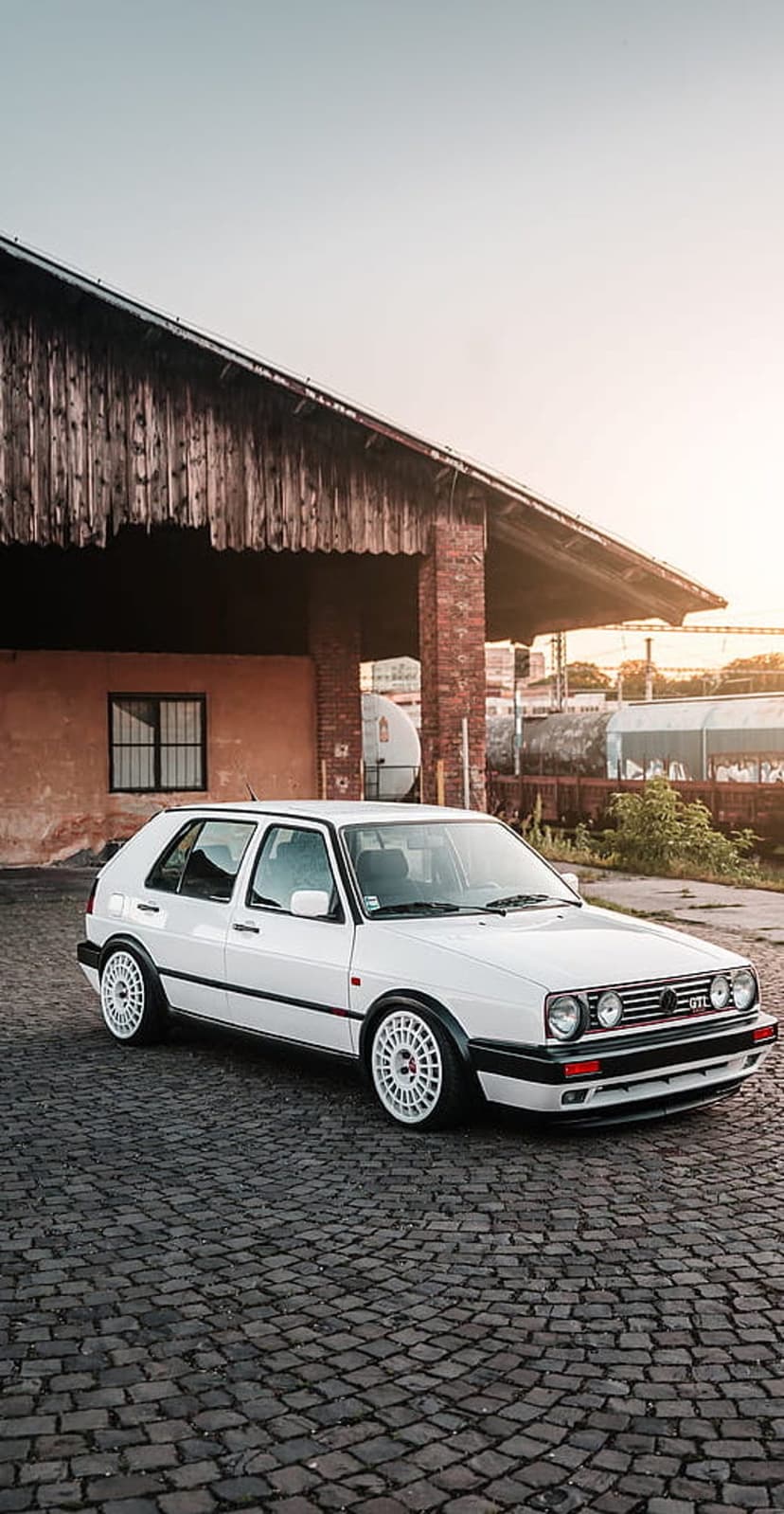 White Volkswagen Golf MK2 GTI parked on a cobblestone street in front of a rustic brick and wood building at sunset.