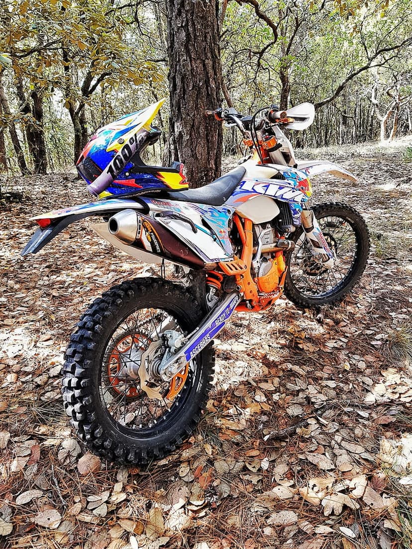 Orange dirt bike with a multi-colored helmet parked on a forest floor covered in golden autumn leaves.