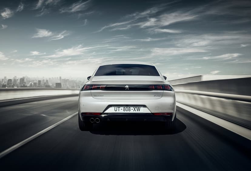 White Peugeot sedan driving on a highway bridge with glowing red taillights and a city skyline background.
