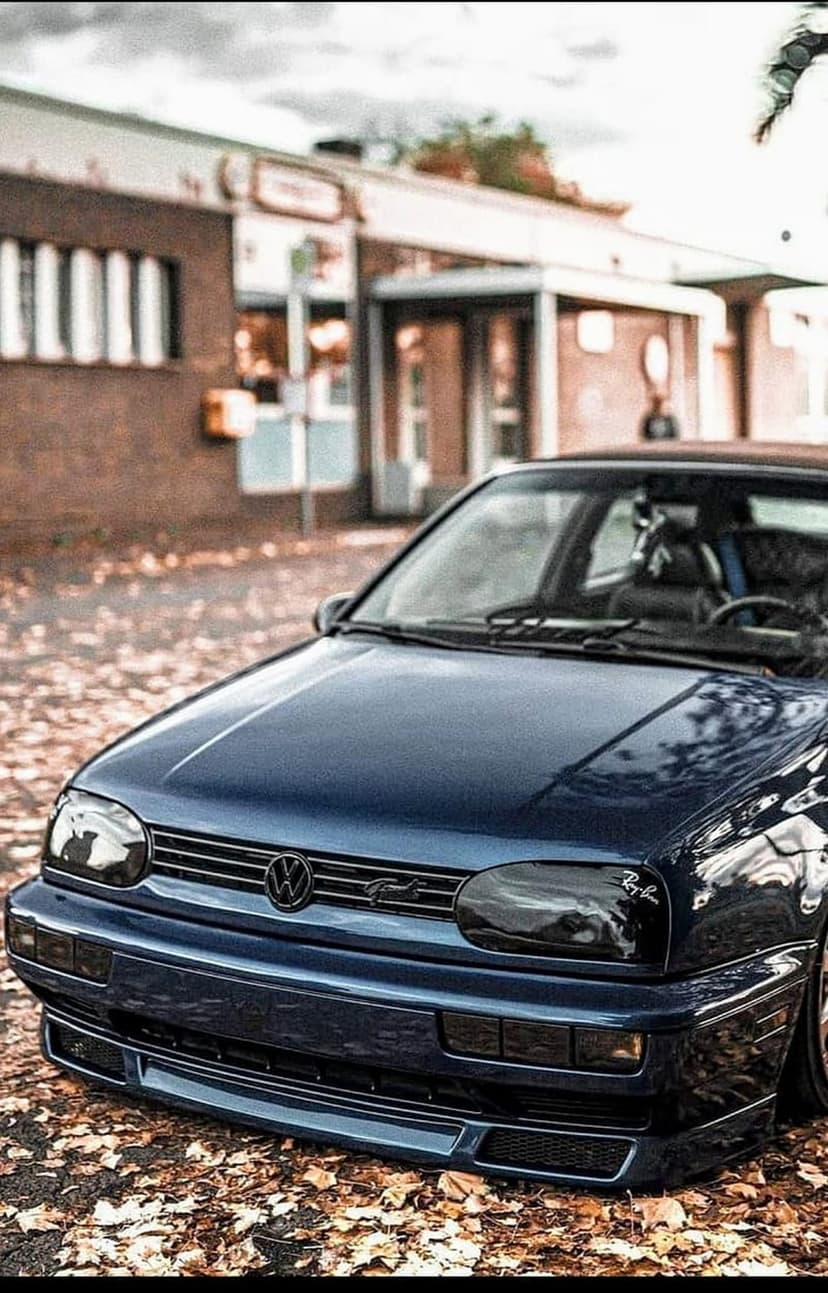 Dark blue Volkswagen Golf GTI mk3 front end on a street with orange autumn leaves in front of a brick building.