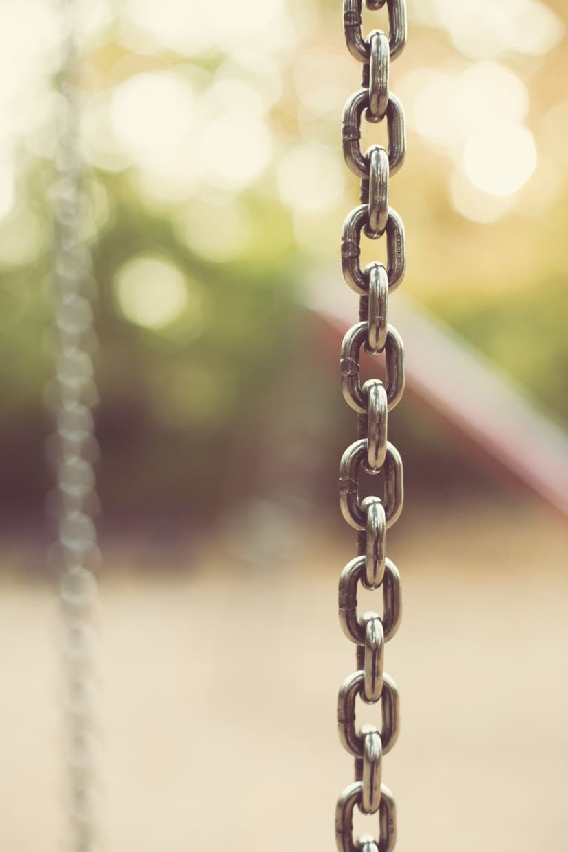 Interlocking silver metal chain links hang vertically against a soft green bokeh background with dappled sunlight.