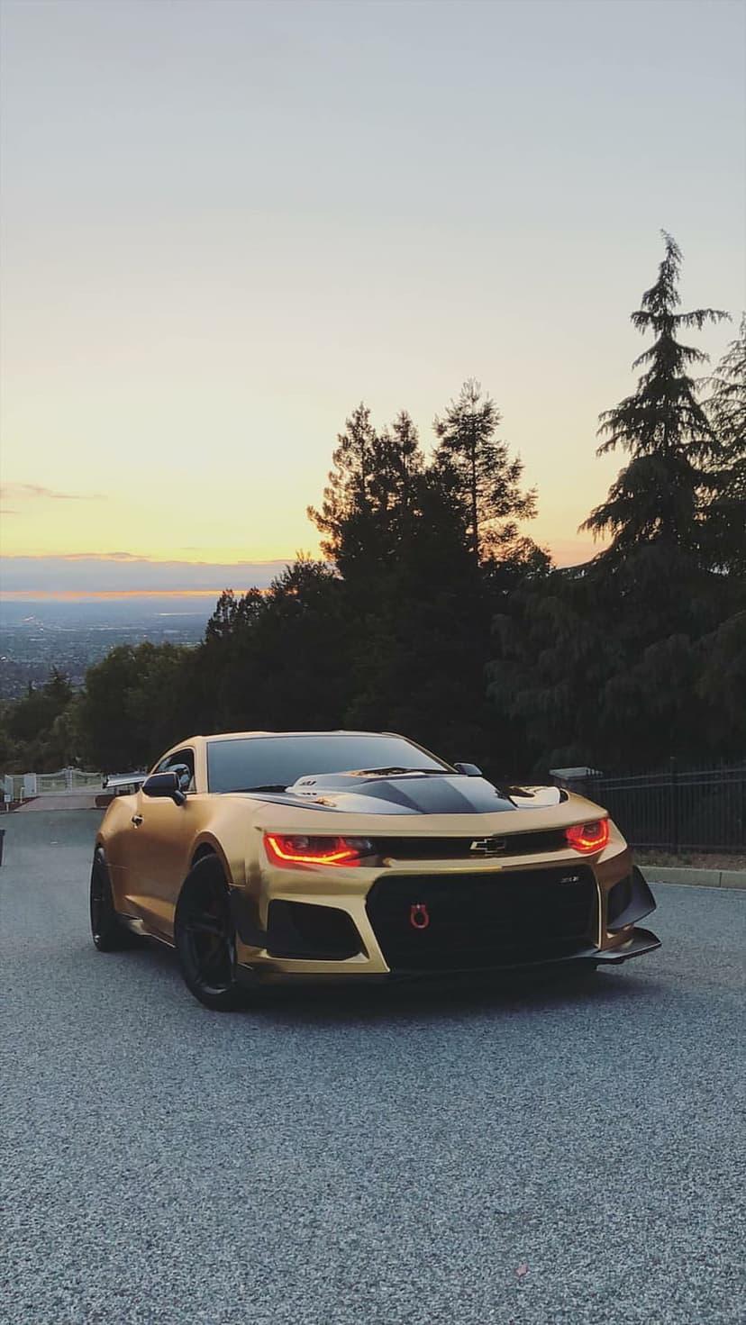 Golden Chevrolet Camaro with red glowing headlights parked on a road overlooking a city at sunset with tall trees.