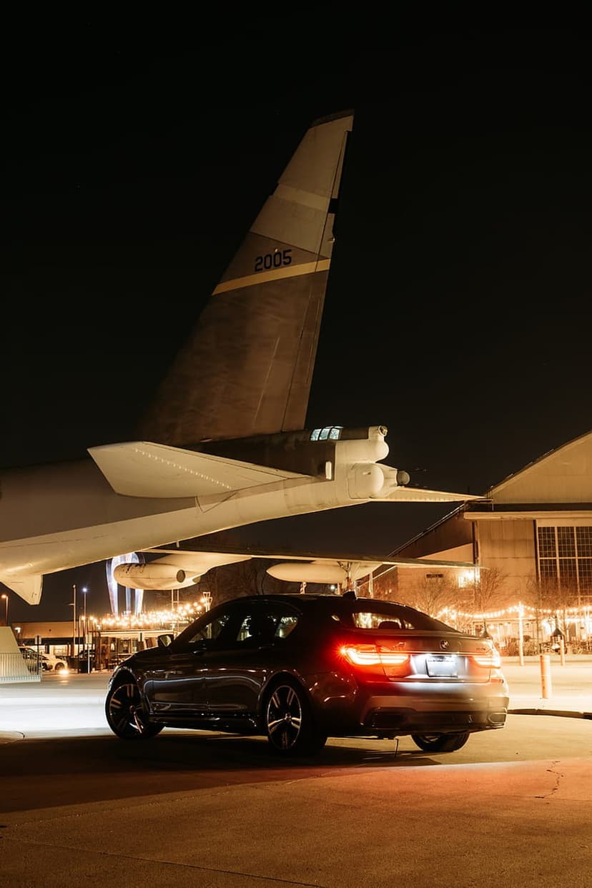 Luxury sedan with glowing red taillights parked on wet asphalt under the wing of a vintage airplane at night.