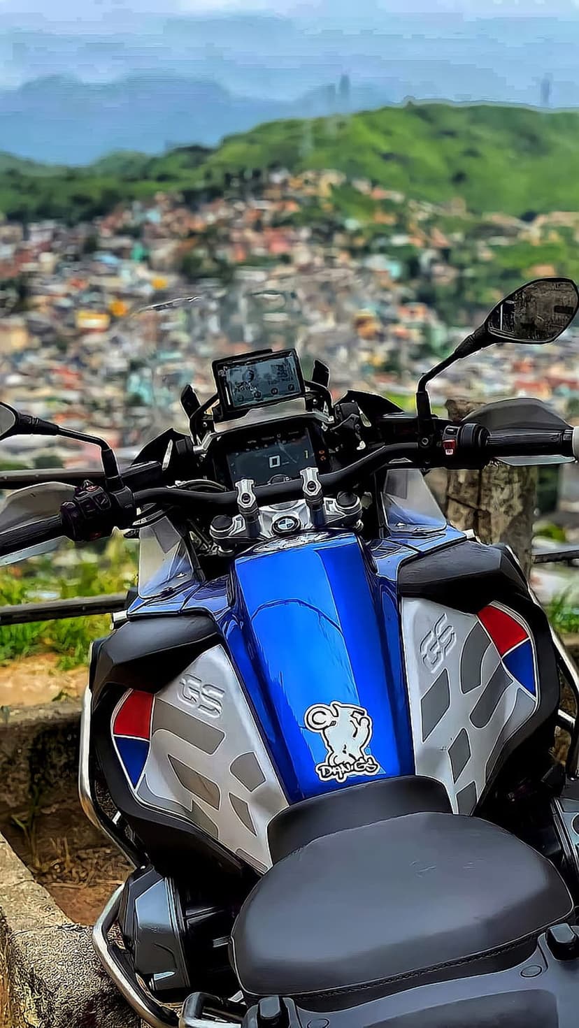 Blue and silver adventure motorcycle handlebars overlook a hazy city valley nestled between green mountains.