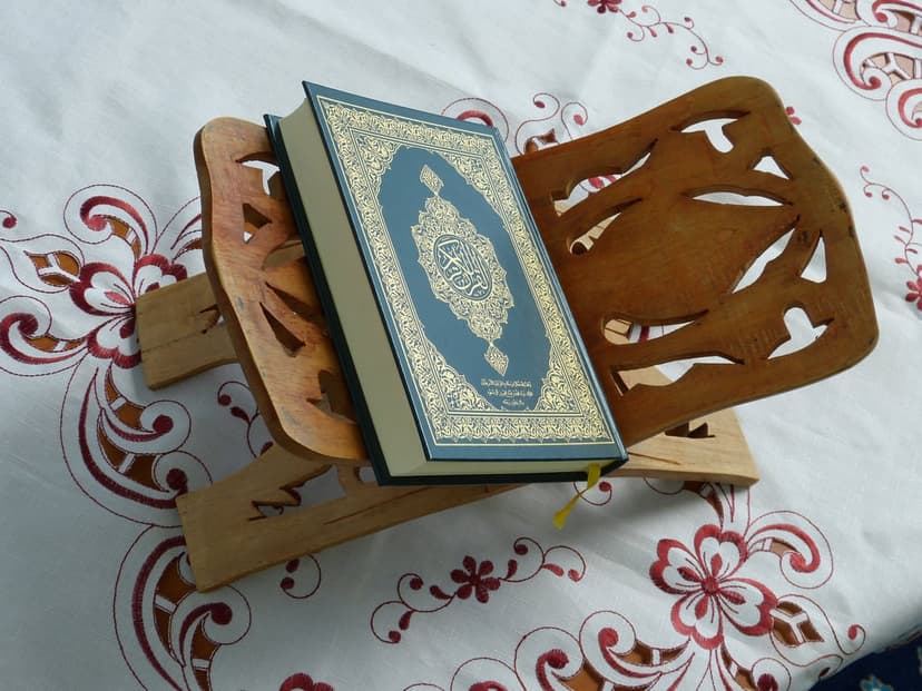 Quran with a blue and gold cover sits on a carved wooden stand atop a white floral embroidered tablecloth.