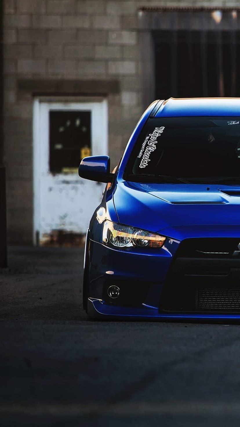 Blue Mitsubishi Evo X with custom decals parked in front of a weathered red brick wall and white door.