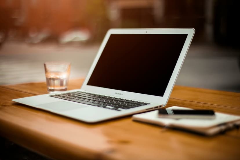 Silver laptop and smartphone on a notebook sit next to a glass of water on a sunlit wooden cafe table.