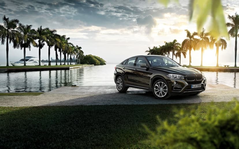 Luxury dark brown BMW X6 parked on a pier with palm trees and a yacht under a warm golden sunset sky.