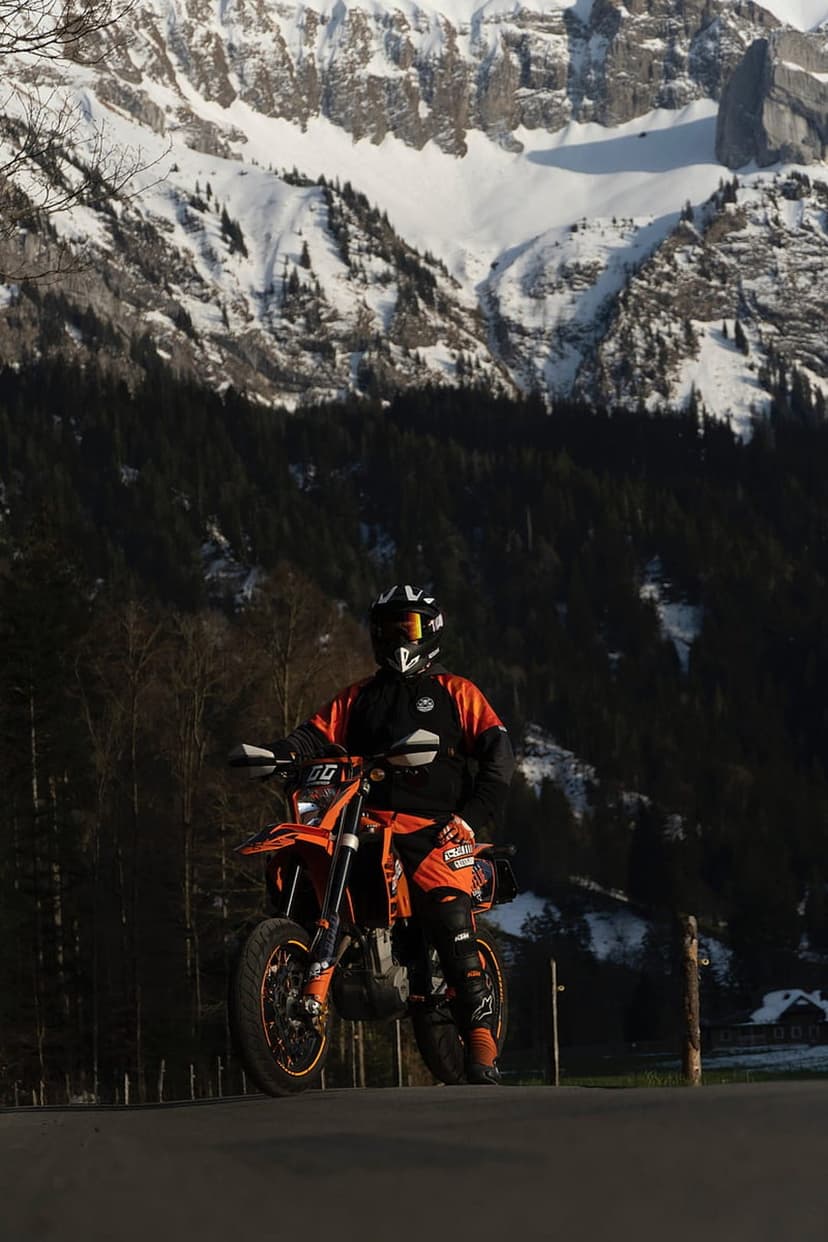 Motorcyclist in orange gear sits on a matching bike with snow-covered mountains and pine trees in the background.