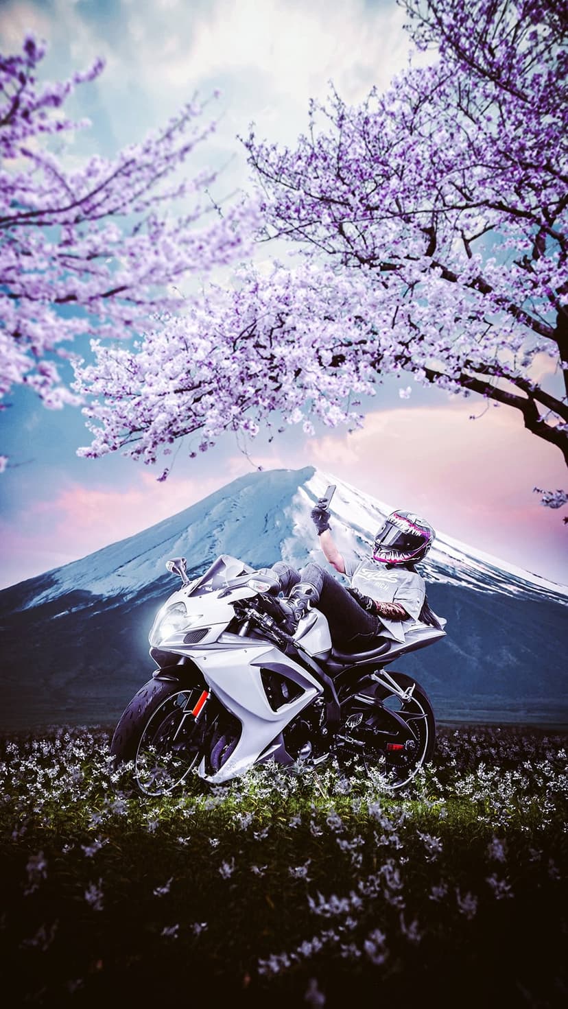 Motorcyclist in a helmet sits on a white sportbike taking a selfie near Mount Fuji and pink cherry blossoms.