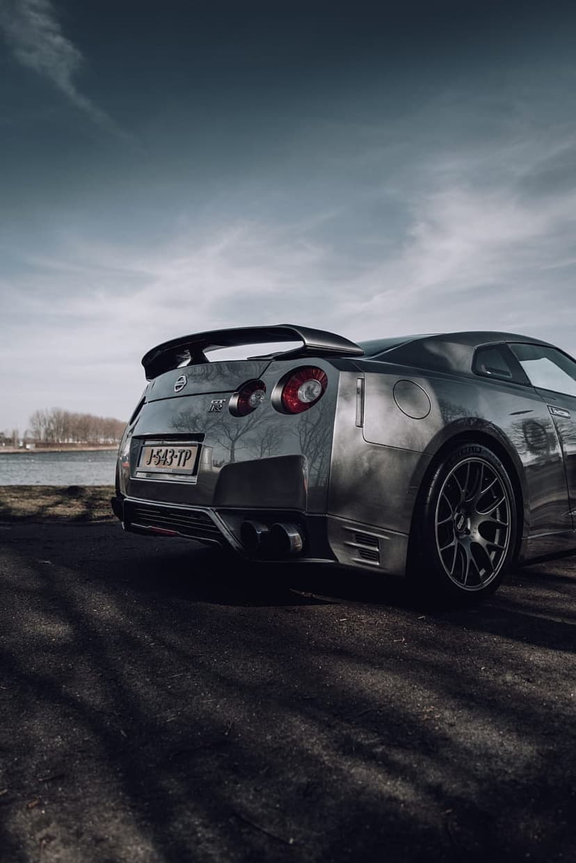 Silver Nissan GT-R rear view with quad taillights and a large spoiler against a dramatic cloudy sky and water.