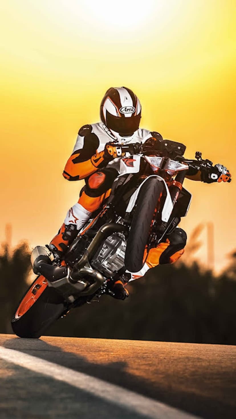 Motorcyclist in full gear performs a wheelie on a KTM bike against a vibrant, orange-hued sunset sky.