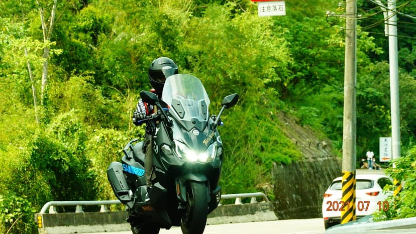 Motorcyclist in a helmet riding a dark touring bike along a winding road surrounded by dense green forest trees.