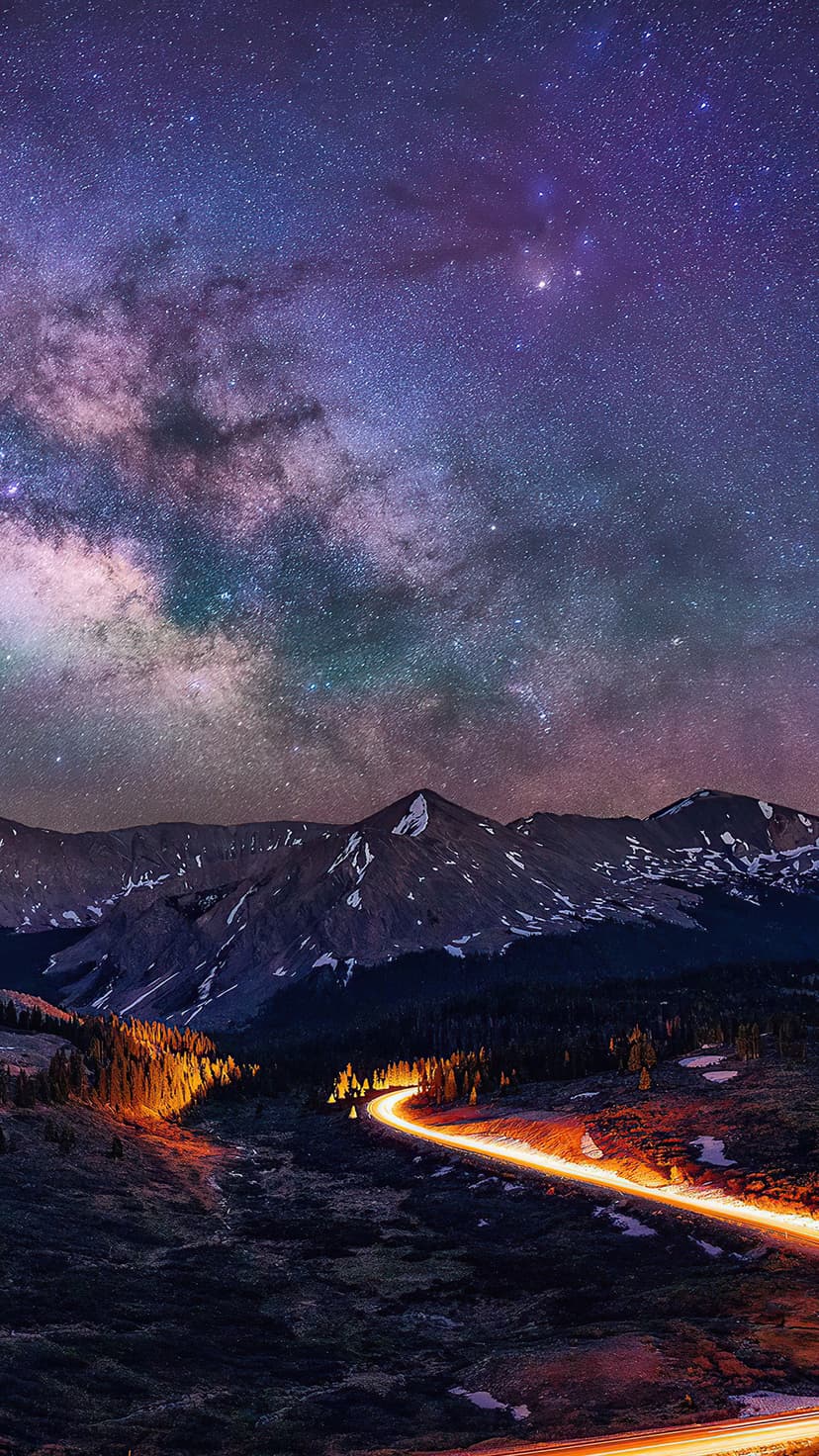 Milky Way galaxy arches over snowy mountain peaks and a winding road with glowing orange vehicle light trails.