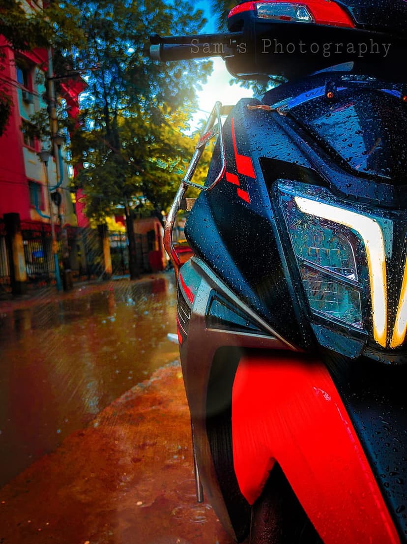Red scooter parked on a reflective wet street with water droplets, city buildings, and lush green trees behind it.