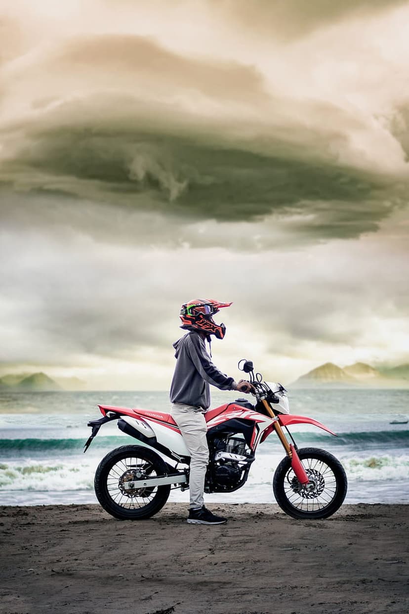 Motorcyclist in a red and white helmet stands by a dirt bike on a sandy beach under dark, stormy clouds.