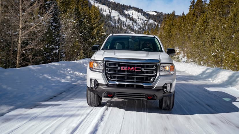 White GMC truck driving on a snowy mountain road lined with pine trees under a clear blue sky in winter.