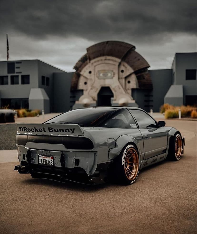 Grey Nissan 240SX Rocket Bunny build with bronze wheels parked by a circular window under a dark cloudy sky.