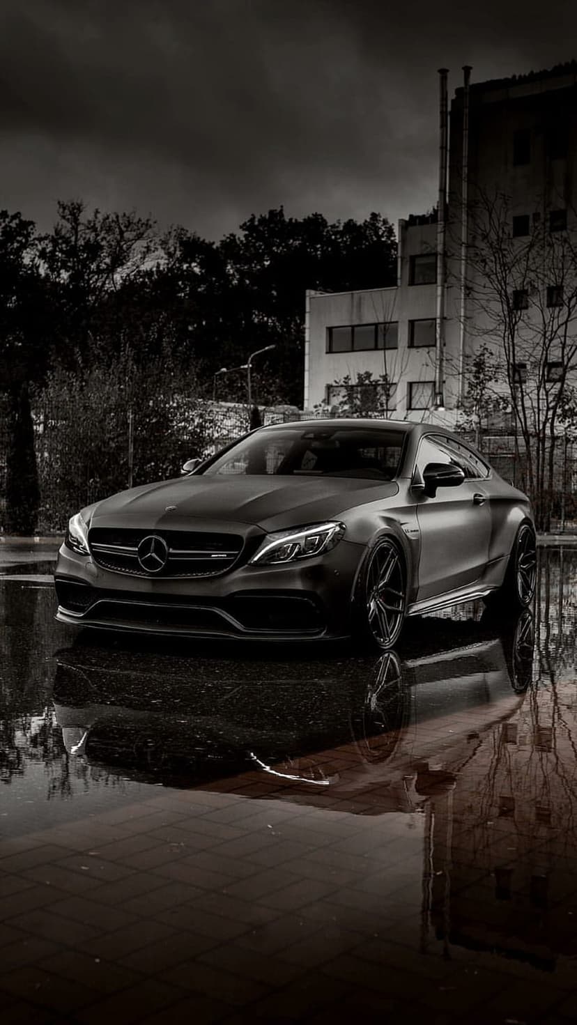 Matte black Mercedes-AMG C63s Coupe parked on wet asphalt reflecting city lights and a dark rainy sky.