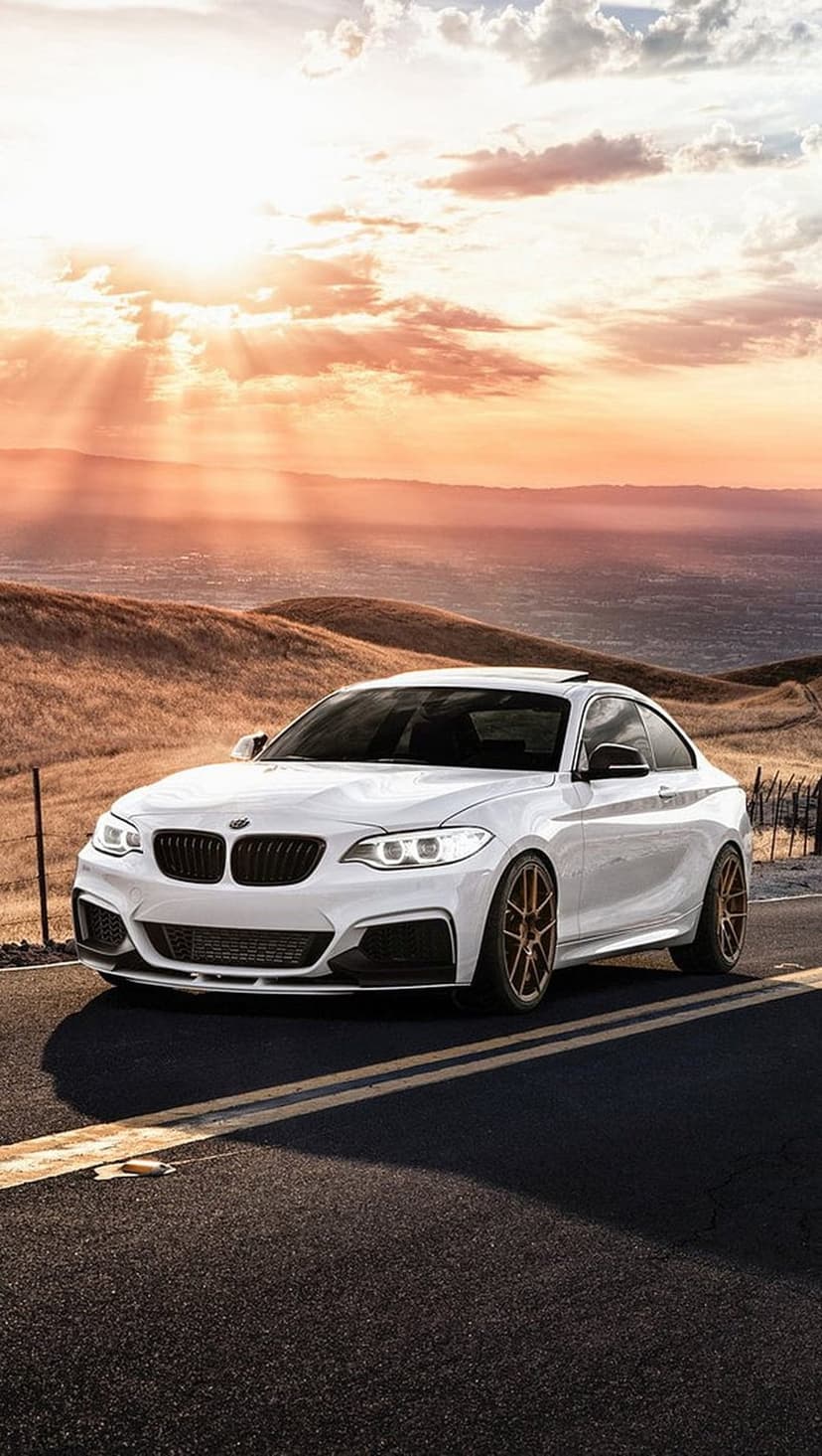 White BMW M2 with bronze rims parked on a scenic road during a golden hour sunset over rolling hills.