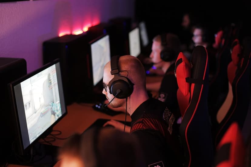 Man with glasses and a black gaming headset plays an FPS game in a dark room with red and purple lights.
