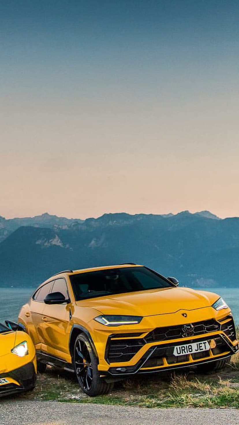 Two yellow Lamborghini Urus SUVs parked on a rocky shore with distant mountains and a calm lake background.