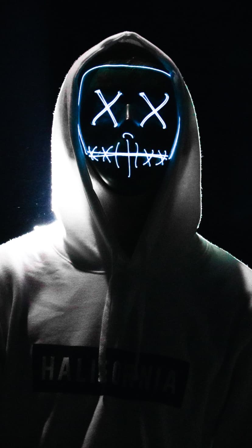 Hooded person wearing a glowing neon mask with X-shaped eyes and a sinister grin against a black background.