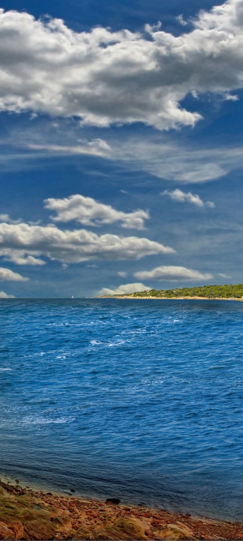 Emerald green coastline meets a deep blue sea under towering white clouds with a small sailboat on the horizon.