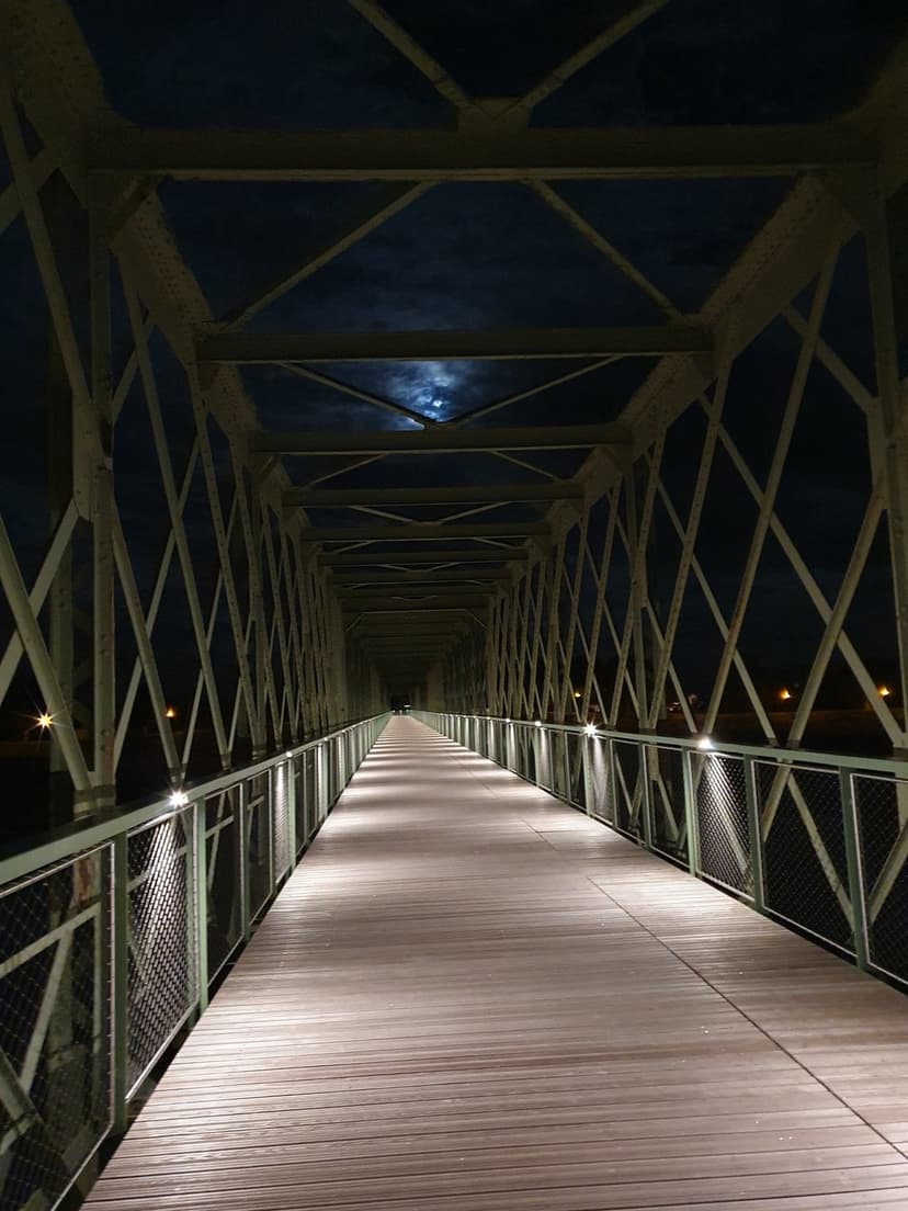 Pedestrian bridge walkway glows under warm lights and a bright moon through steel girders in the night sky.