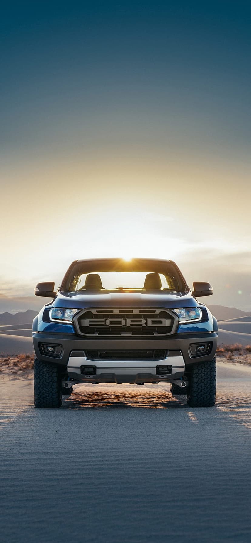 Ford Ranger Raptor truck with glowing headlights driving through sandy desert dunes during a golden sunset.