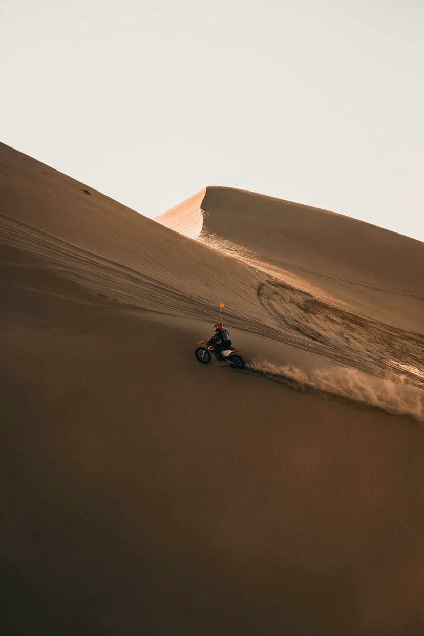 Motorcyclist in an orange jacket rides a dirt bike down a massive sand dune, kicking up dust in a vast desert.