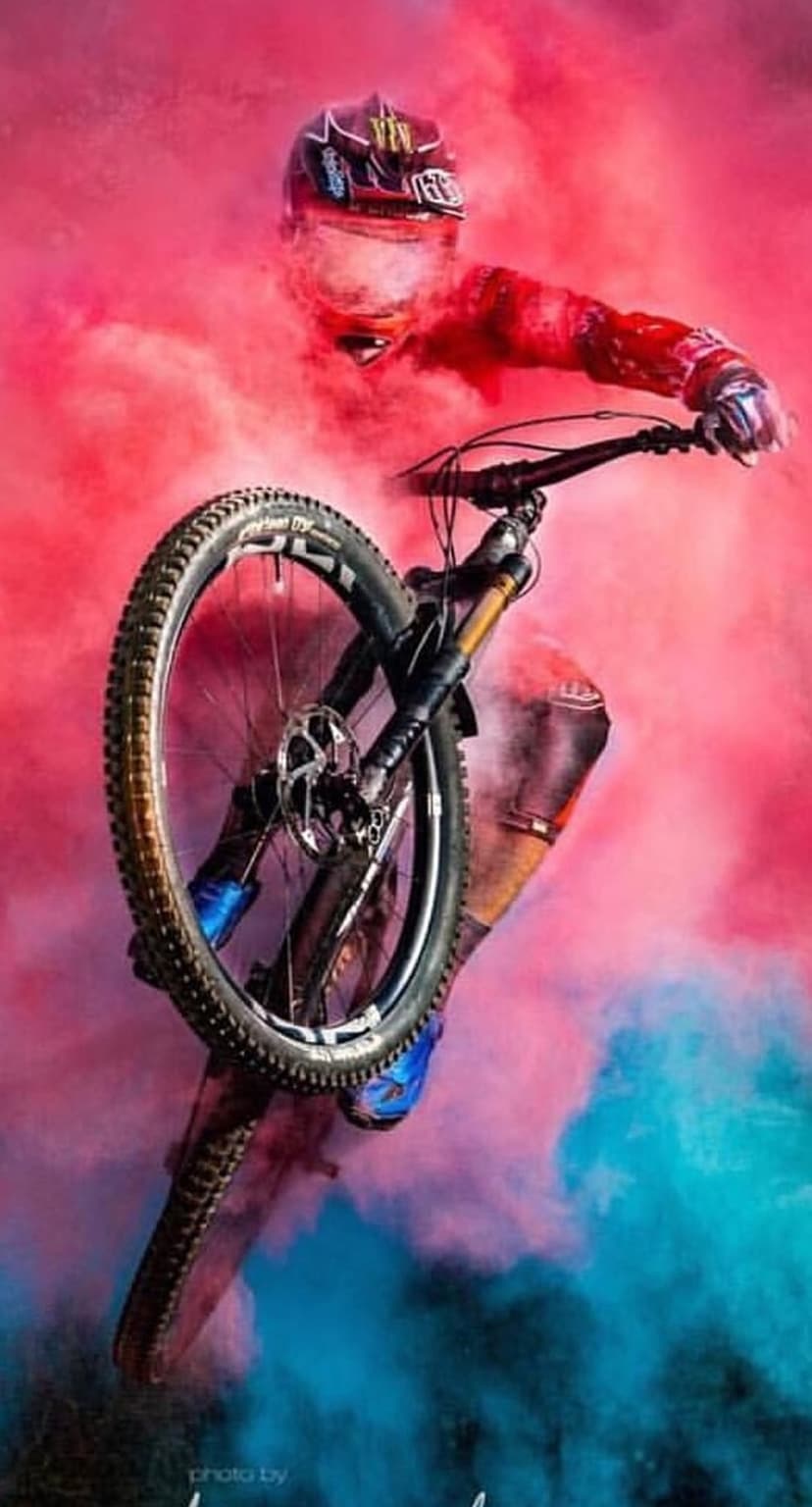Mountain biker in a red jersey mid-jump surrounded by thick plumes of vibrant pink and blue smoke clouds.