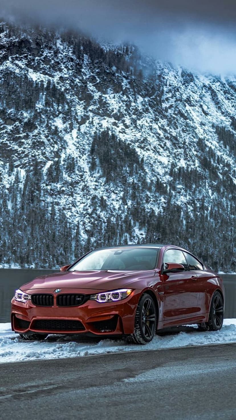 Red BMW M4 coupe parked on a snow-covered mountain road with glowing headlights and jagged peaks in the distance.