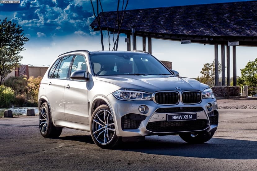 Silver BMW X5 M SUV parked on asphalt with a rustic wooden structure and dramatic blue sky clouds background.