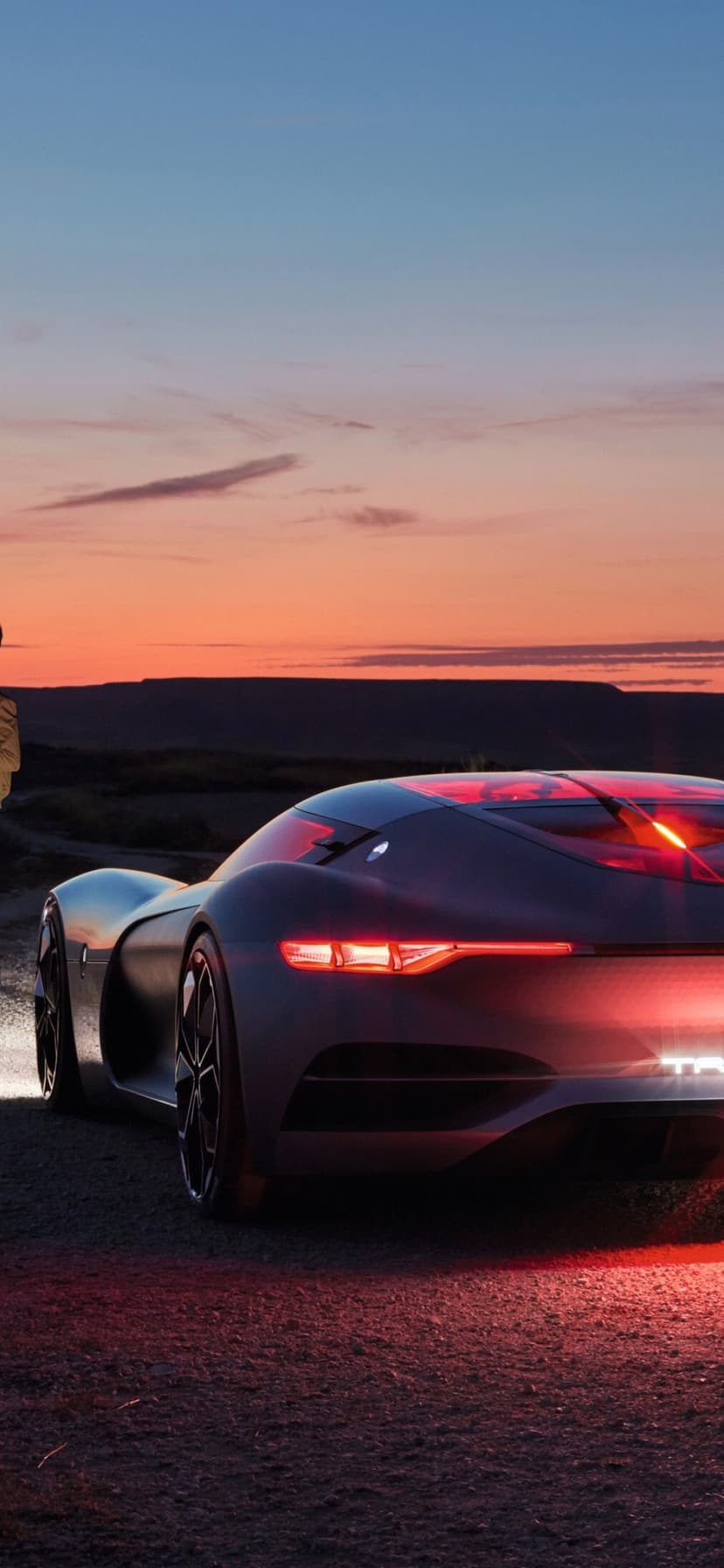 Matte grey concept car with red tail lights parked on a dirt road under an orange and blue sunset sky.