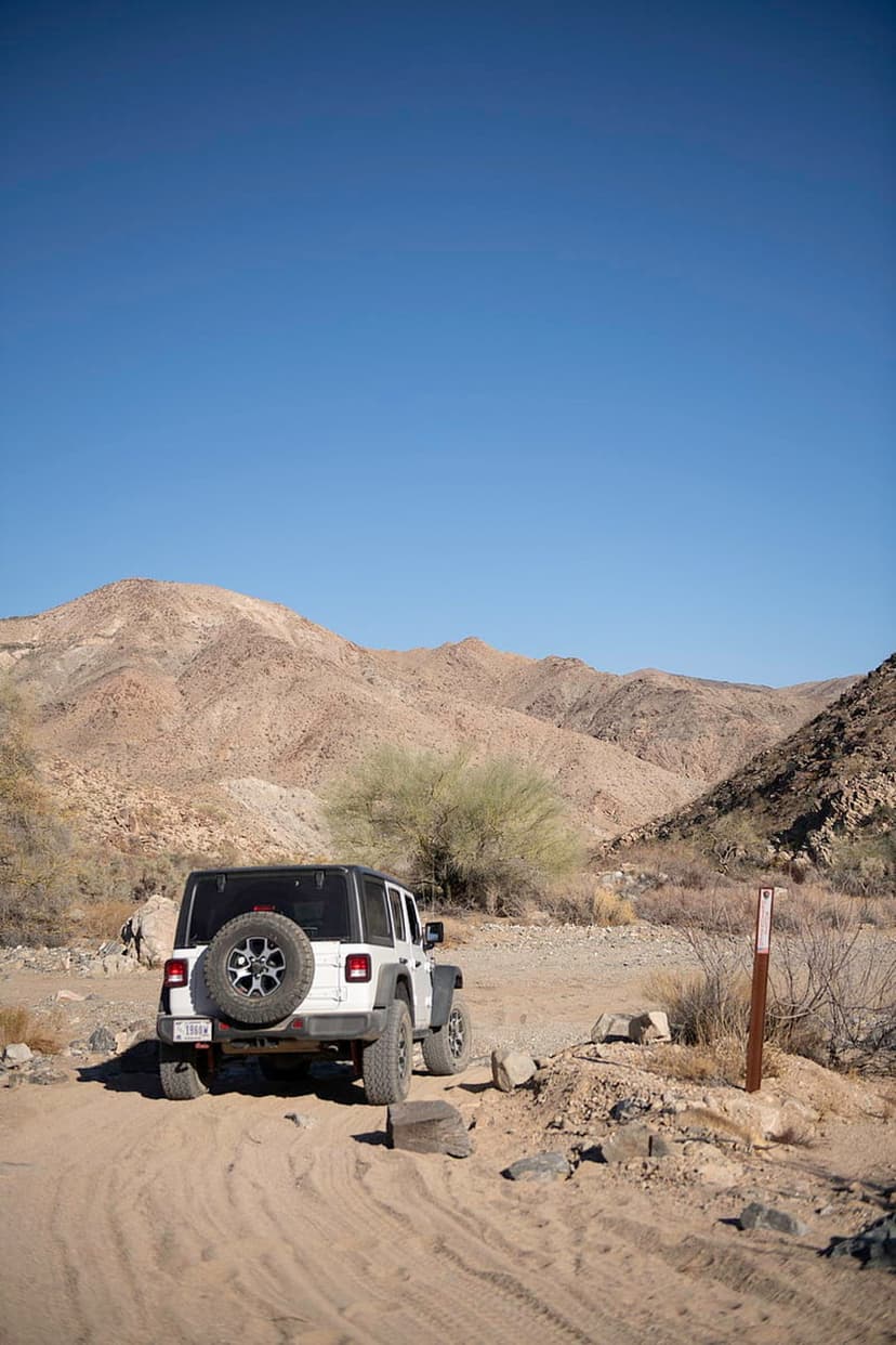 White Jeep Wrangler with a spare tire driving on a sandy trail toward rugged mountains under a clear blue sky.