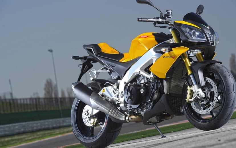 Yellow Aprilia Tuono V4 sportbike on a racetrack with gold forks, silver frame, and a clear blue sky background.
