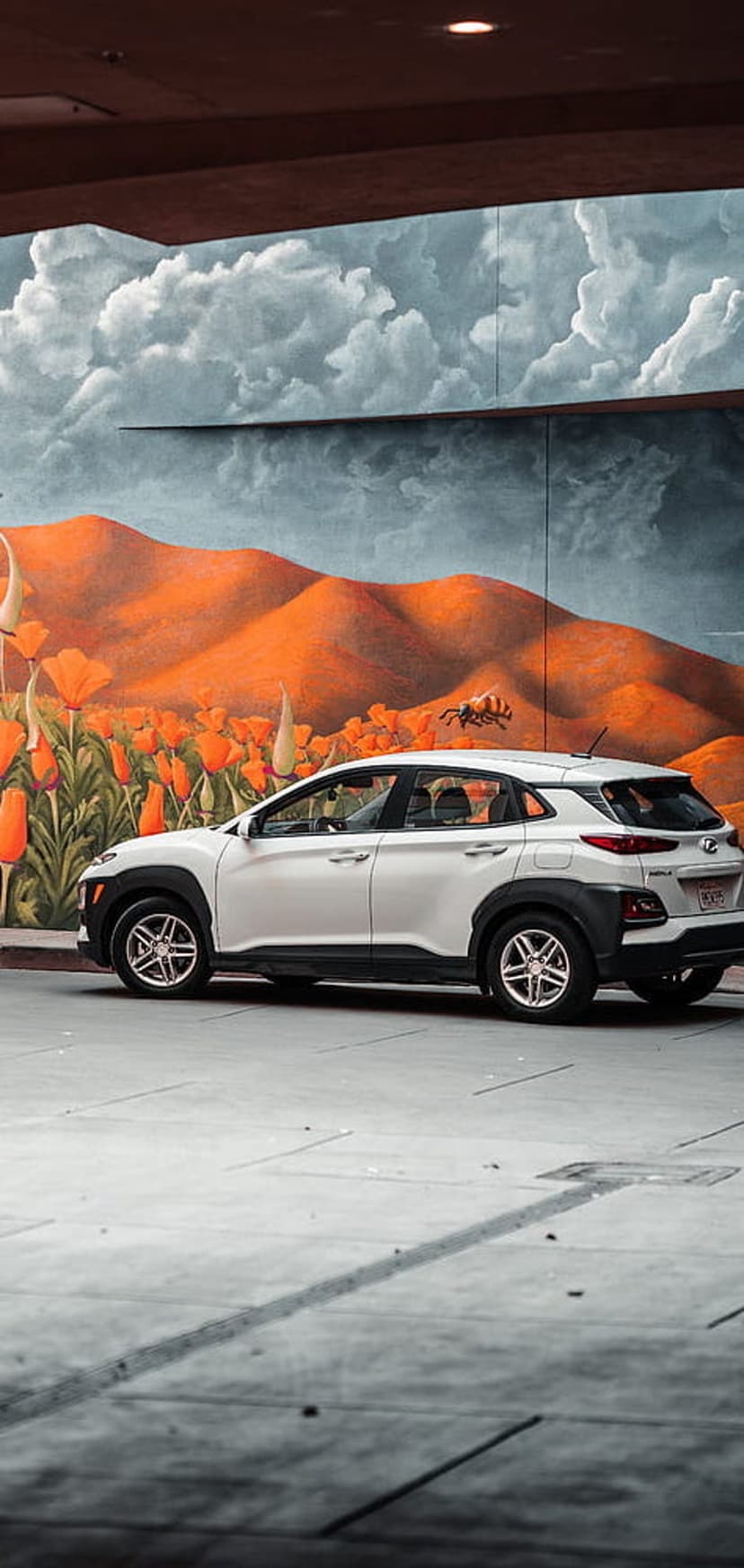 White Hyundai Kona parked in front of a colorful wall painting featuring orange hills and large red flowers.
