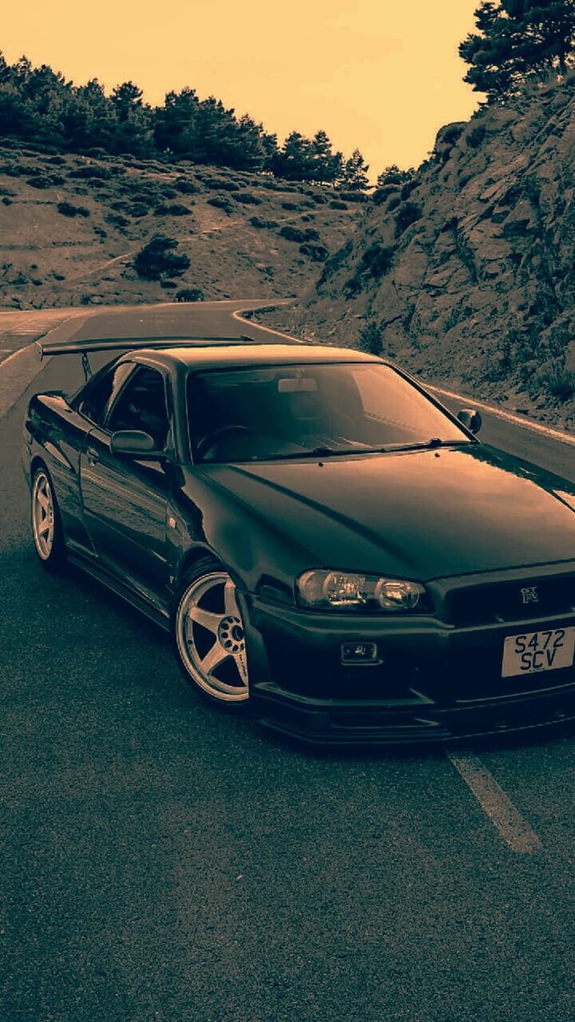 Dark Nissan Skyline R34 GT-R driving on a winding mountain road under a warm sunset with pine trees nearby.