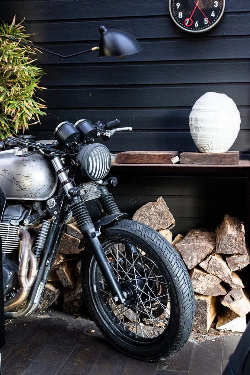 Vintage motorcycle parked next to a firewood pile and white vase against a dark wood wall with a wall clock.