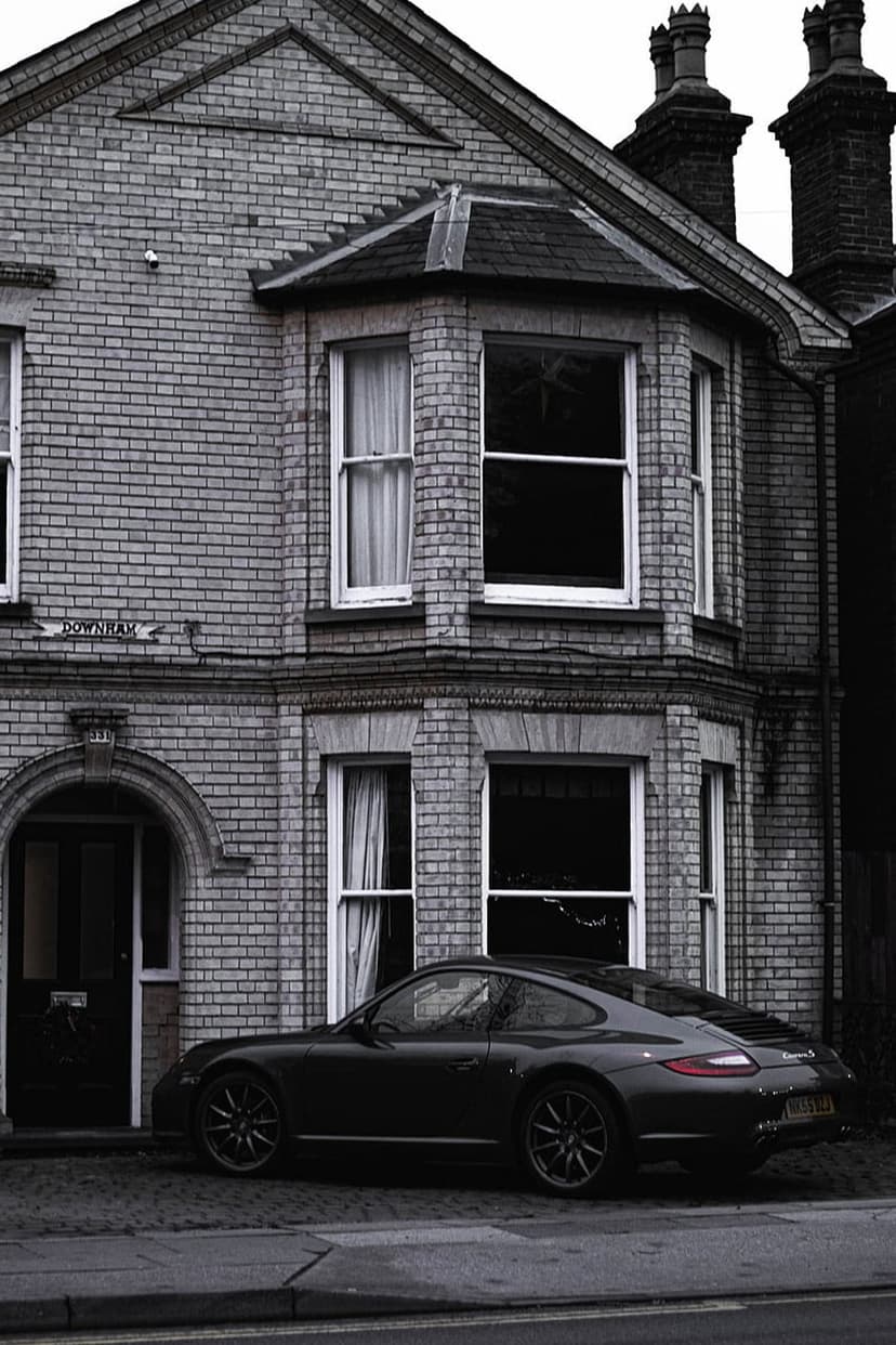 Black Porsche 911 parked on a cobblestone street in front of a two-story brick house with a bay window.