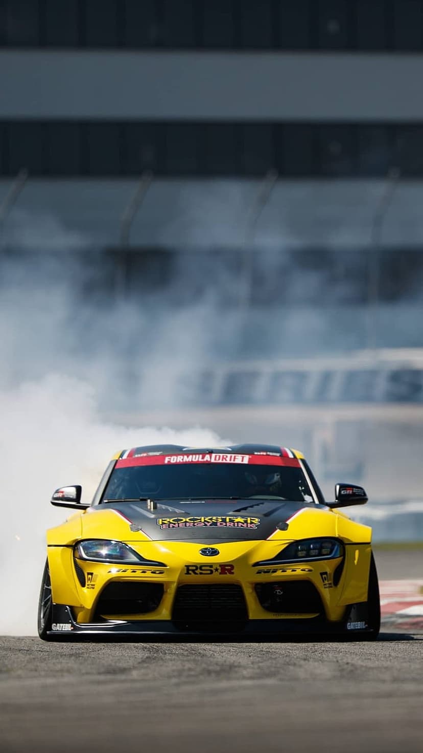 Yellow Toyota Supra drifting on an asphalt track with thick white tire smoke and Rockstar Energy branding.