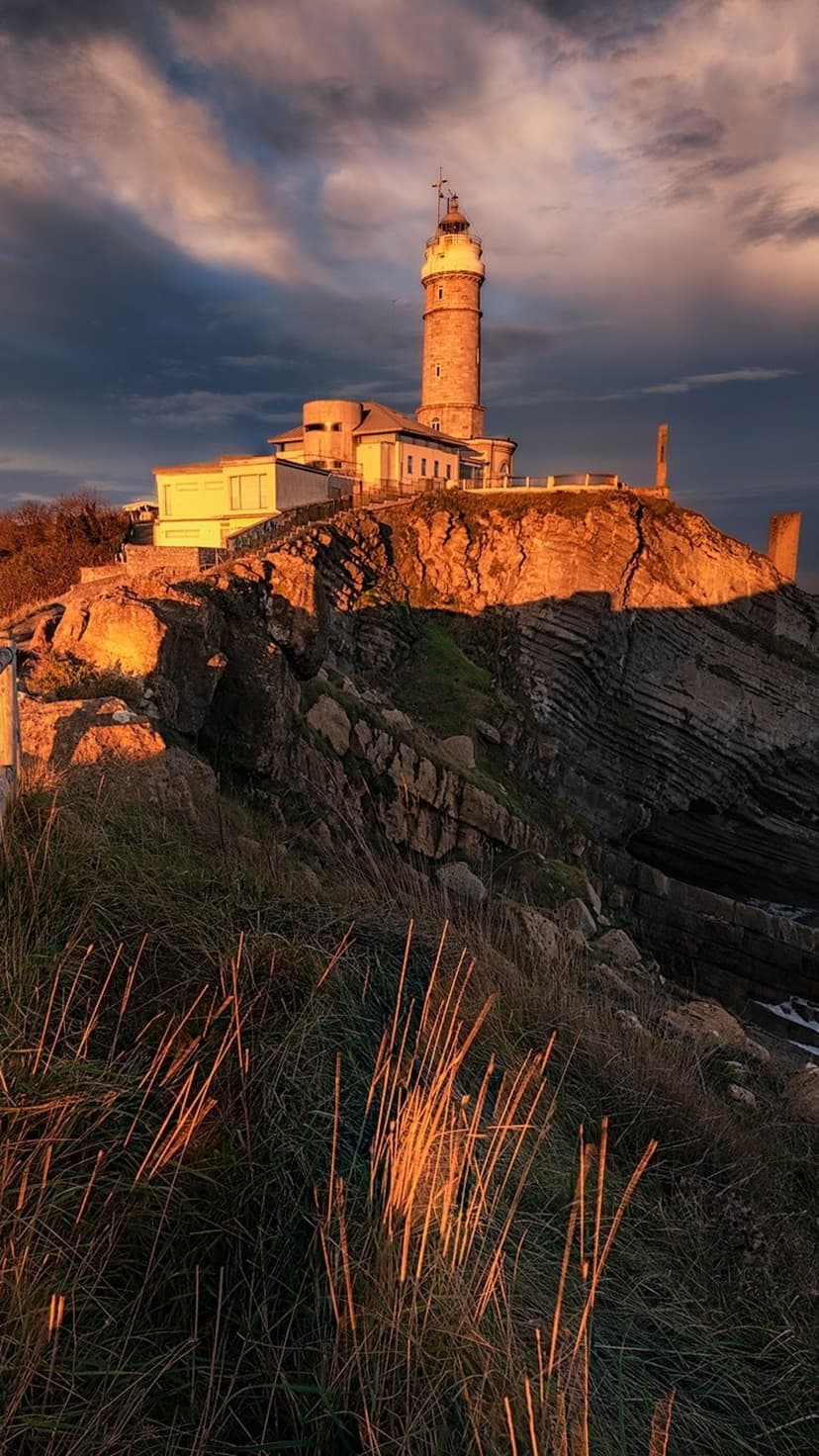 Tall stone lighthouse atop a steep cliff overlooks the vast ocean under a dramatic golden and orange sunset sky.