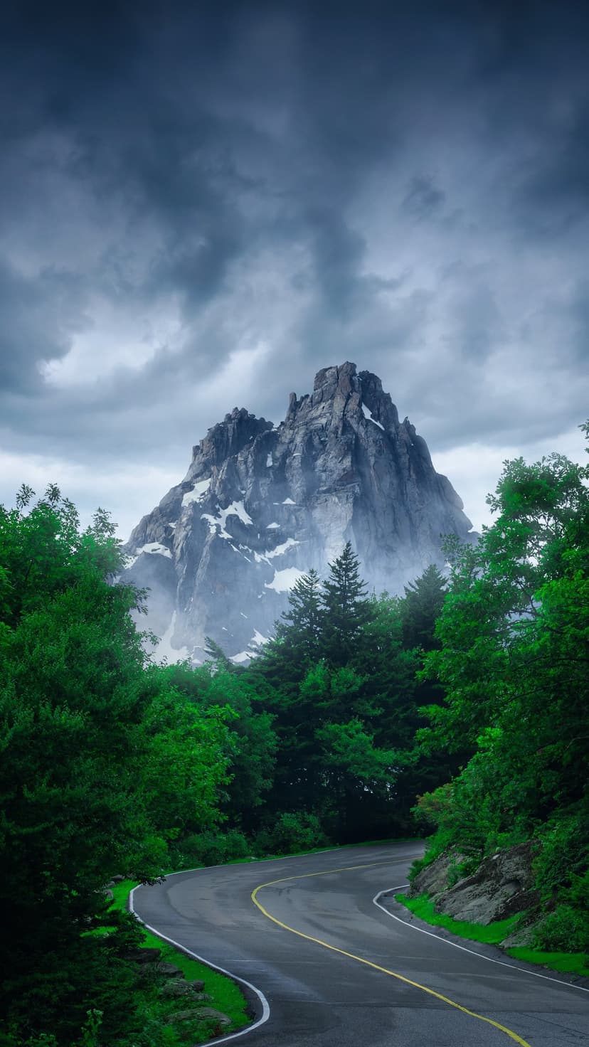 Winding asphalt road curves through green forests toward a rugged, snow-dusted peak under heavy storm clouds.